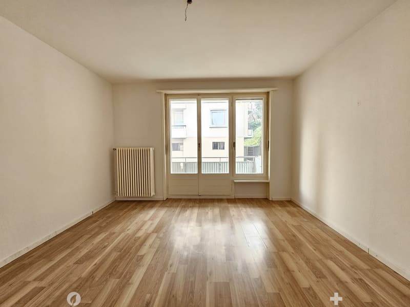 Spacious living room with hardwood floors, large windows, and a radiator. The room appears to be empty, ready for furnishing and decorating.