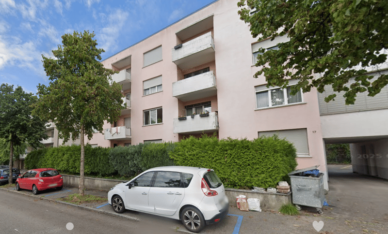 3-story apartment building with balconies, surrounded by trees and greenery, with parked cars in the foreground