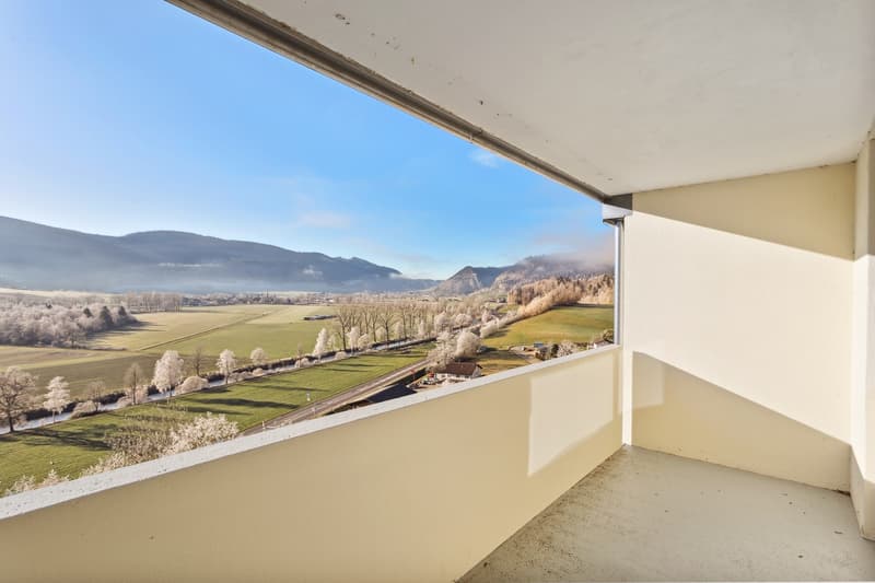 The image shows a balcony or terrace with a panoramic view of a snowy, mountainous landscape. The balcony has a concrete floor and is surrounded by a railing. The view outside includes snow-covered trees, fields, and distant mountains under a blue sky.