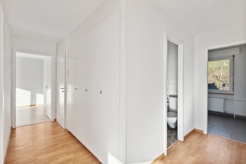 The image shows a hallway with white walls, wooden floors, and built-in closets along one side. There is a doorway leading to another room visible at the end of the hallway.