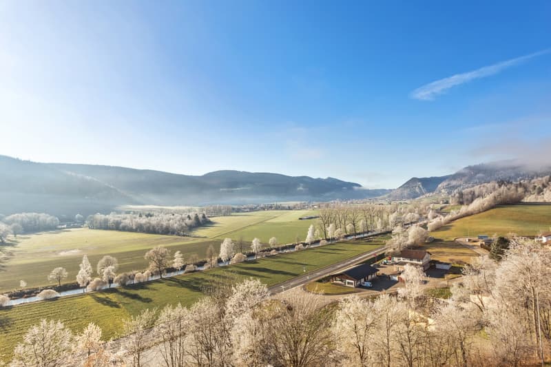 Scenic rural landscape with rolling hills, snow-covered trees, and a road winding through the valley. The image shows a picturesque countryside setting with a clear blue sky and distant mountains in the background.