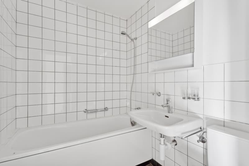 Tiled bathroom with a white bathtub, sink, and shower head. The walls are covered in white square tiles arranged in a grid pattern.