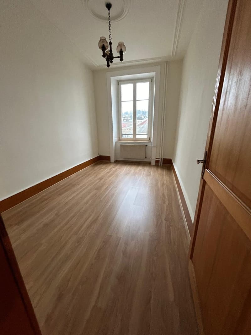 Spacious bedroom with hardwood flooring, white walls, and a large window providing a view of the surrounding area. The room features a chandelier lighting fixture.