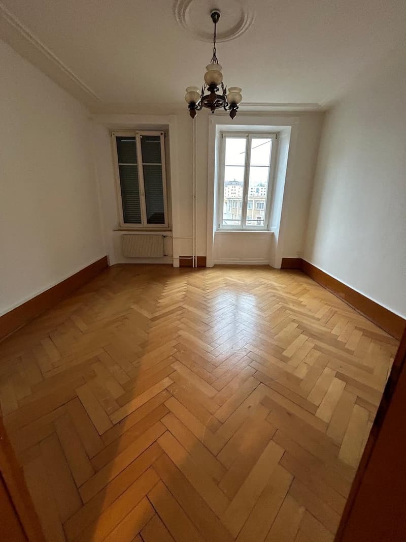 This image shows a spacious room with hardwood flooring in a herringbone pattern. The room has large windows that provide natural light, and a decorative chandelier hanging from the ceiling. The walls are painted a neutral color, creating a clean and mini