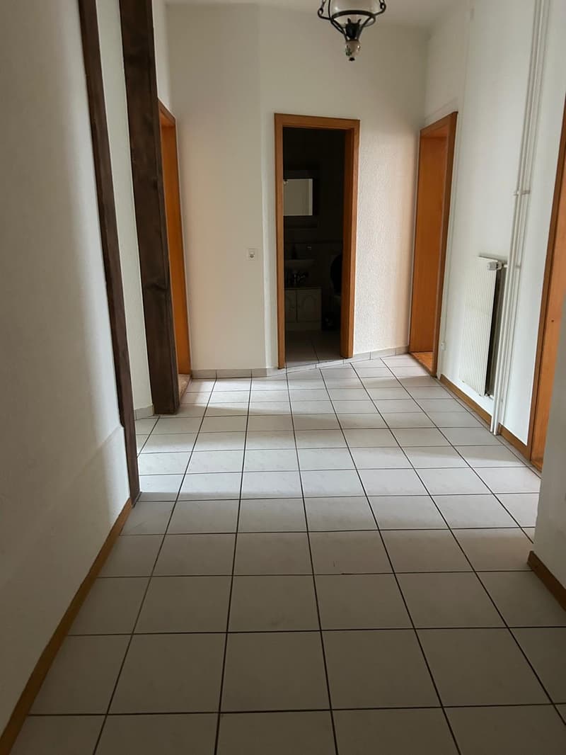 The image shows a hallway with white walls, a tiled floor, and wooden door frames. The hallway appears to be well-lit with a light fixture hanging from the ceiling.