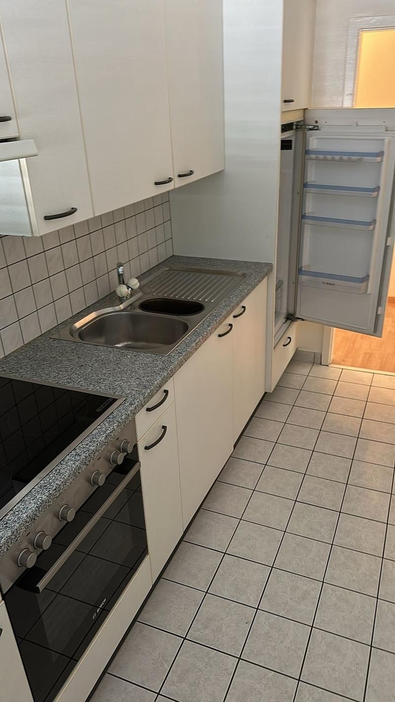 Compact kitchen with white cabinets, granite countertop, stainless steel sink, built-in dishwasher, and a refrigerator. The floor is tiled with a neutral color scheme.