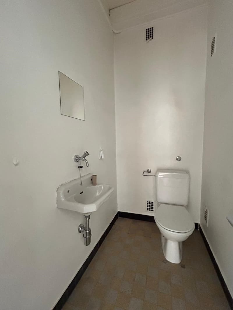 Small bathroom with a white sink, toilet, and tiled floor. The walls are painted white and there is a small mirror on the wall.