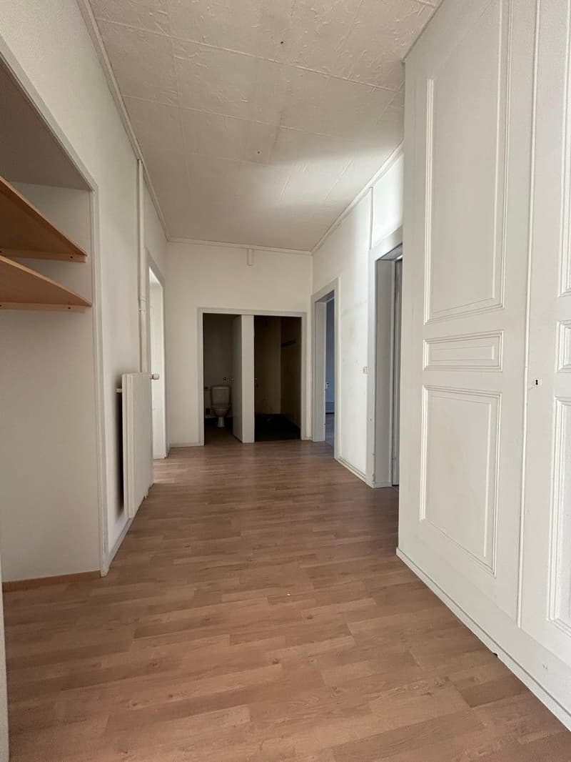 The image shows a long, narrow hallway with white walls and a wooden floor. There are several doors leading to other rooms along the hallway, and some shelving units can be seen on the walls.