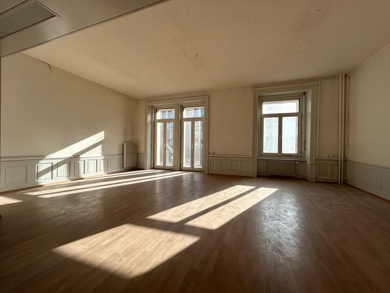 Large, open room with hardwood floors, white walls, and large windows allowing natural light to flood the space. The room appears to be empty, with no furniture or decor visible.