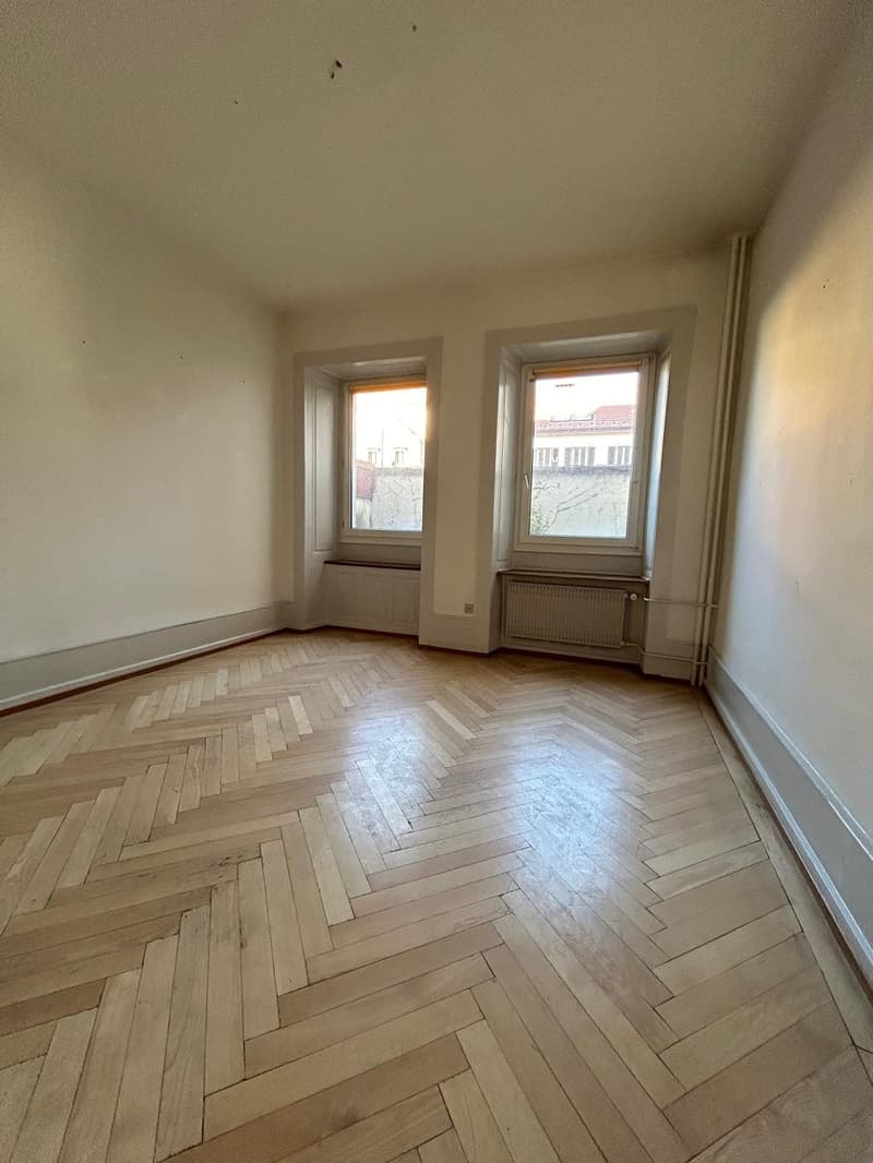 This image shows a room with a hardwood floor in a herringbone pattern. The room has two large windows that provide natural light. The walls are painted a neutral color. The room appears to be empty, with no furniture or decor visible.