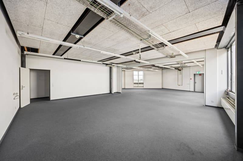Large open office space with concrete ceiling, exposed ductwork, and gray carpeted flooring. The room has a few doors leading to other areas and a window providing natural light.