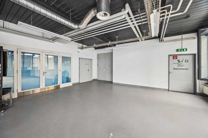 The image shows a large open office space with a high ceiling and exposed ductwork. The walls are mostly white, with some glass partitions and doors. The floor is a concrete or industrial-style flooring. There are signs on the walls indicating this is a s