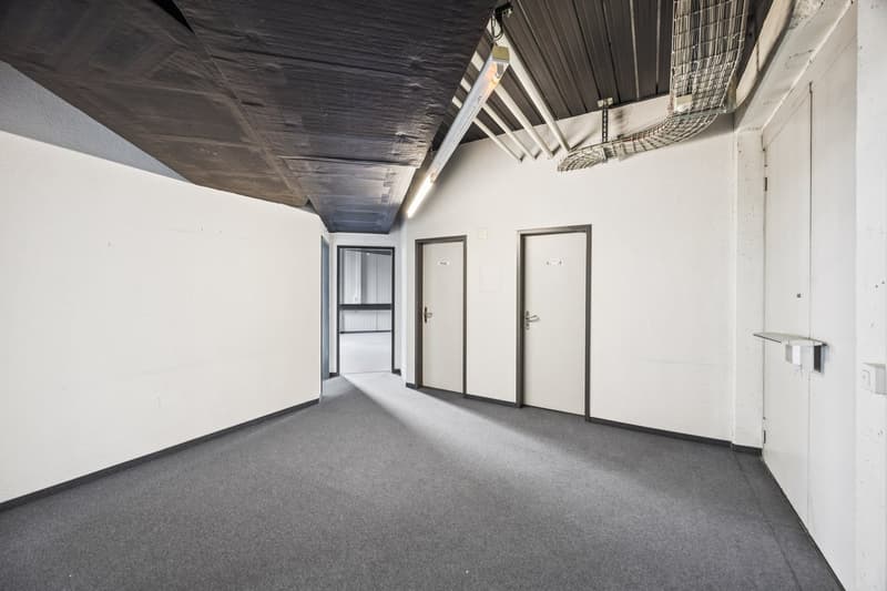 The image shows a large open office space with a high ceiling and exposed ductwork. The walls are white, and the floor is covered in gray carpeting. There are two doors visible, suggesting this could be a multi-room office space. The overall aesthetic app