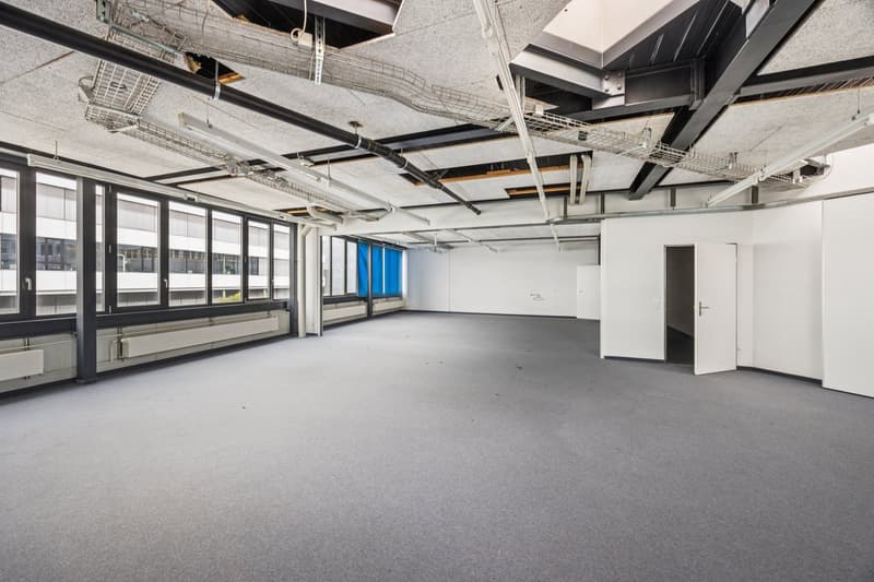 Large open office space with exposed ceiling, concrete floor, and large windows providing natural light. The space appears to be unfinished or under renovation, with visible ductwork and electrical conduits overhead. There is a white door visible in the c