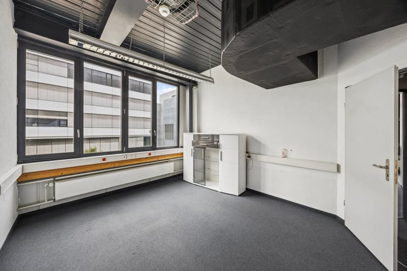 Large open office space with concrete ceiling, black framed windows, gray carpeted floor, and white walls. The room contains a storage cabinet and a small appliance, possibly a refrigerator or microwave.