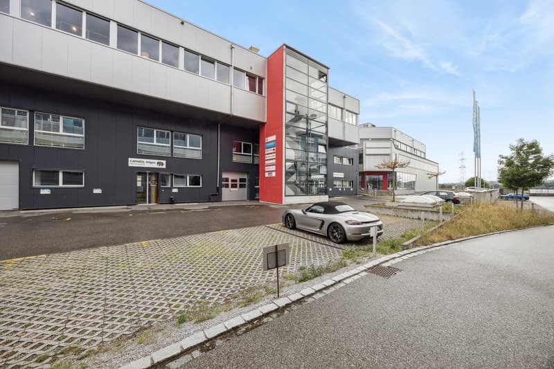 Multi-story commercial building with a modern, industrial design. The building has large windows, a red accent wall, and a parking area in front. There is a car parked in the lot.