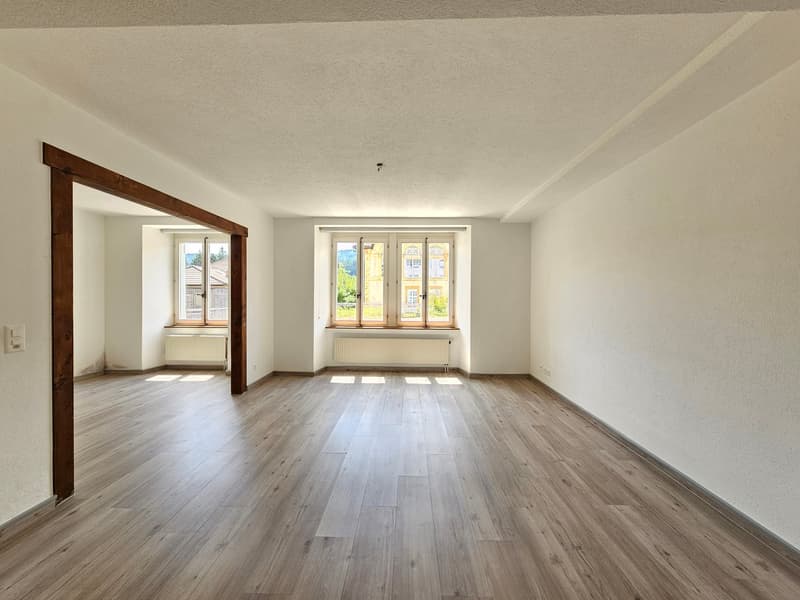 Spacious living room with hardwood flooring, large windows providing natural light, and wooden door frames.