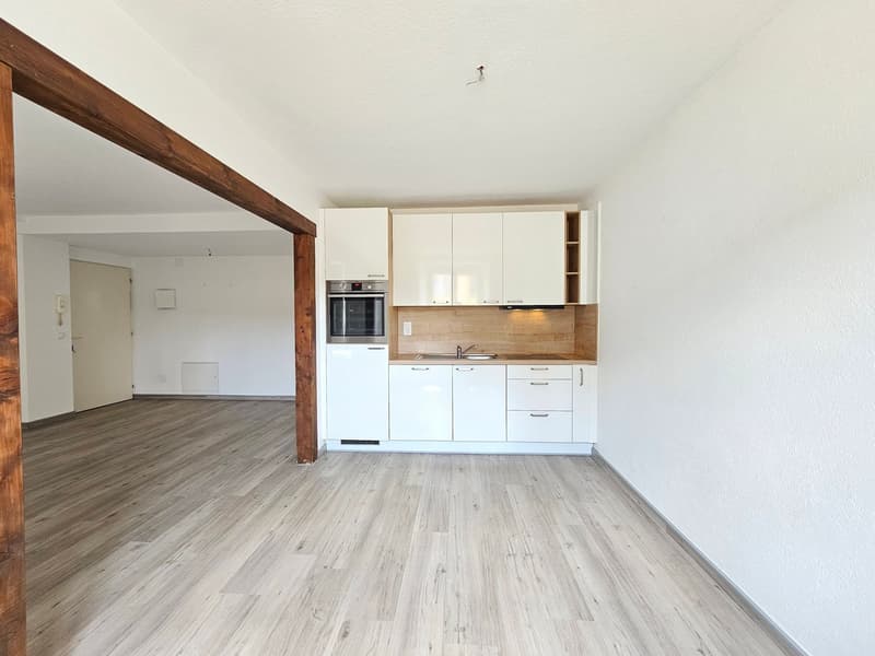 The image shows an open-plan living space with a kitchen area. The kitchen features white cabinets and drawers, a built-in oven, and a wooden backsplash. The flooring throughout the space is a light-colored wood-look laminate. There is a wooden beam visib