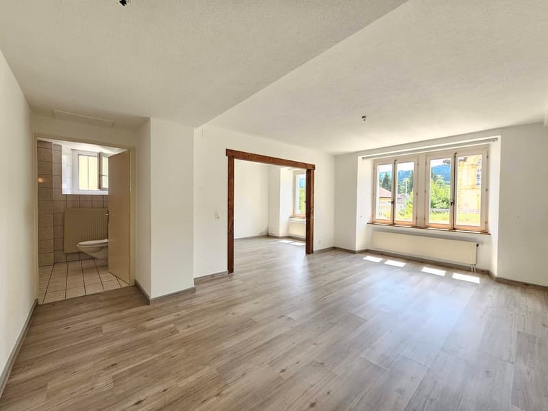 This image shows a spacious and bright apartment interior. The room has a hardwood floor, white walls, and large windows that provide natural light. There is a doorway leading to what appears to be a bathroom. The room is empty, suggesting it is a newly r