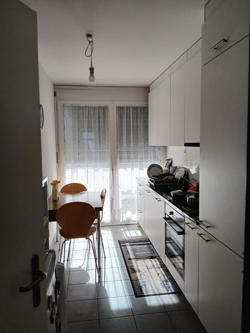 modern kitchen with wooden dining table, two chairs, white cupboards, tiled floor, glass doors leading to balcony, gas stove, oven
