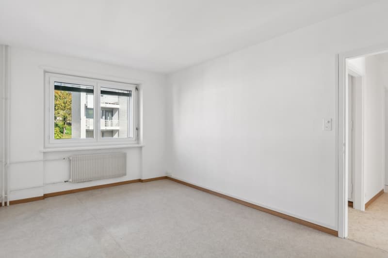 Empty room with white walls, white ceiling, white floor, radiator below the window, door to another room on the right.