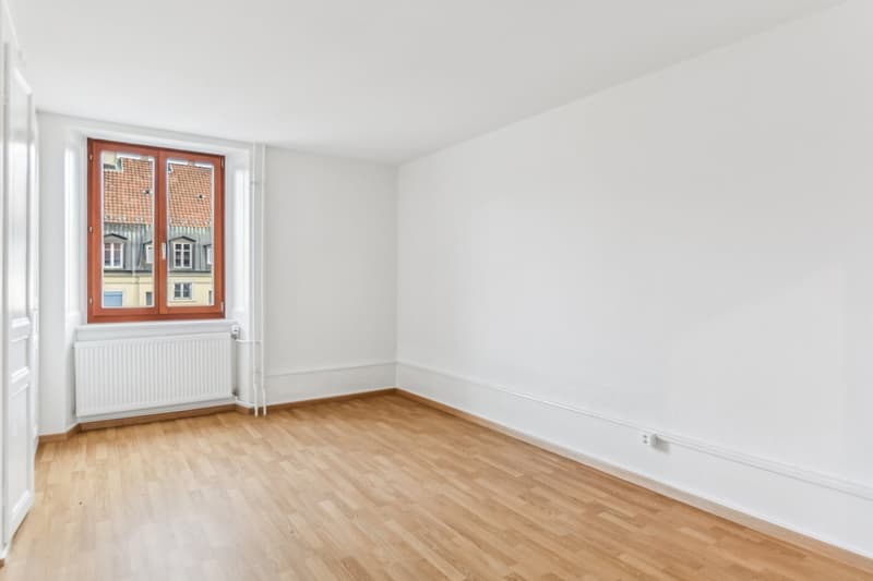 Empty room with wooden floor, white walls, a window, and a radiator.