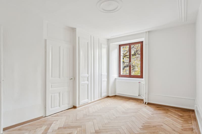 Empty room with white walls, wooden parquet floor, white radiators, double door, and window