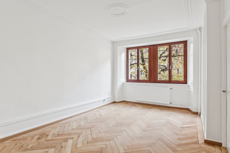 An empty room with wooden herringbone flooring, white walls, and a window with wooden frame