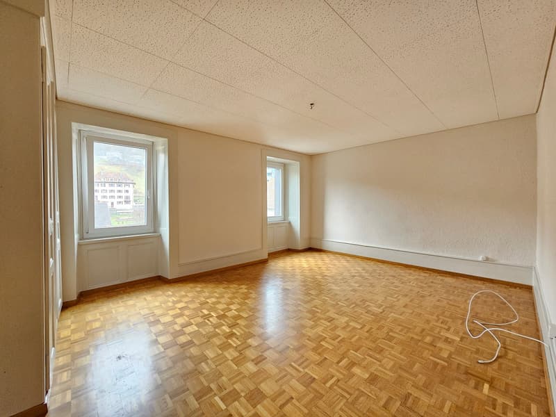 Empty room, wooden parquet flooring, white walls, two windows, ceiling with circular perforations, electrical outlet