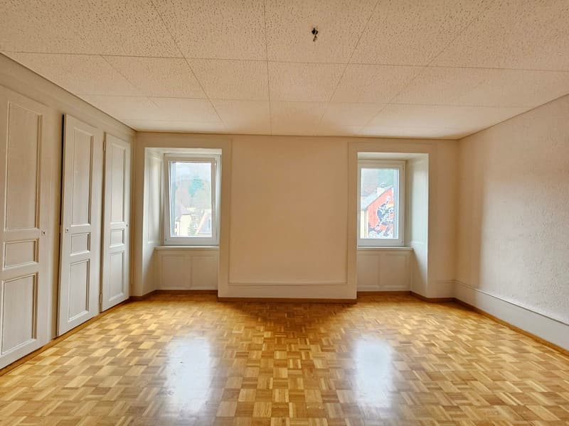 Empty room with wooden parquet flooring, two windows, white walls, and white doors.