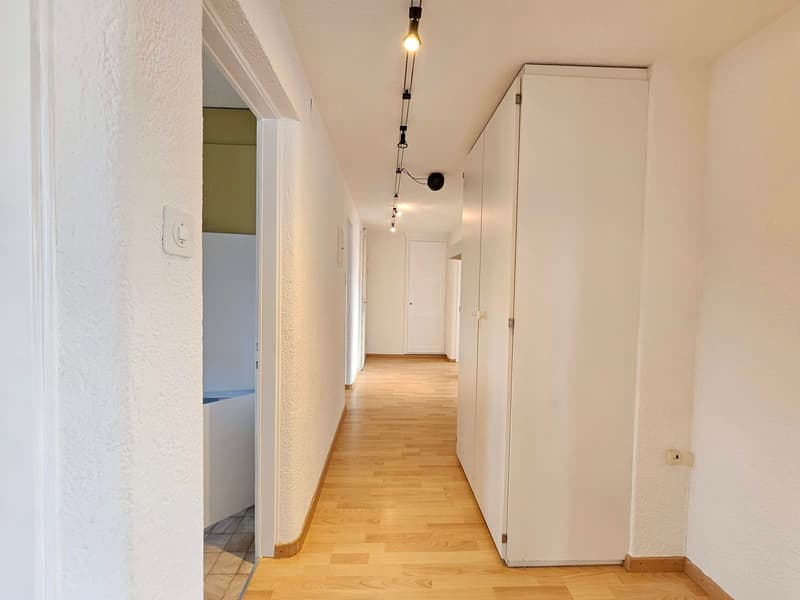 An empty corridor with wooden floors and white walls. There are ceiling lights and an open door at the end.