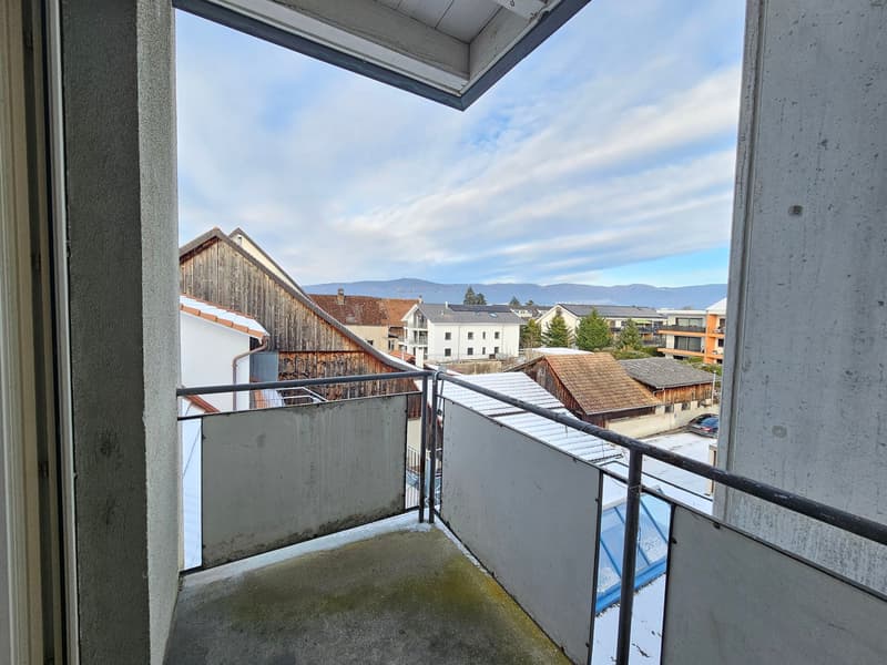 An exterior view from a balcony, overlooking a neighborhood with traditional wooden houses and modern buildings. In the distance, mountains are visible against a blue sky with clouds.