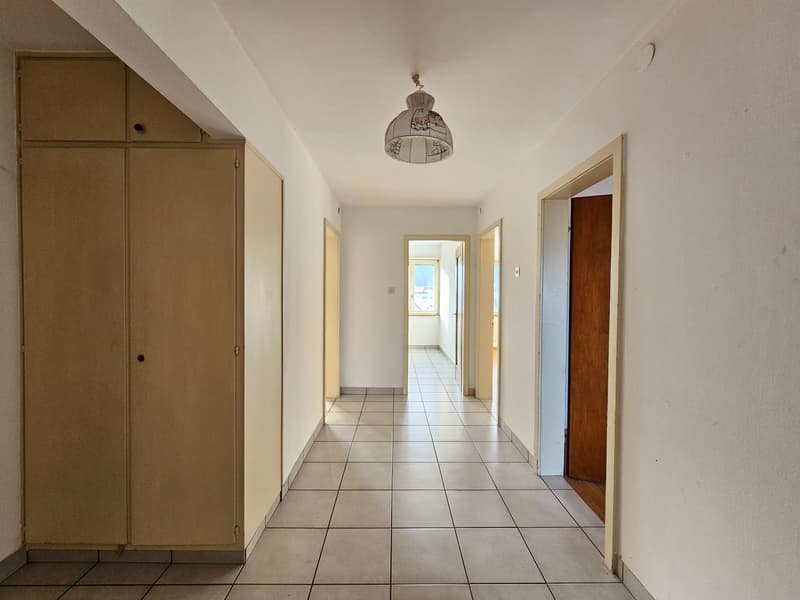 Long hallway with white tiled flooring, wooden cabinets, white walls, single hanging lamp, two doors on the right side, small balcony at the end