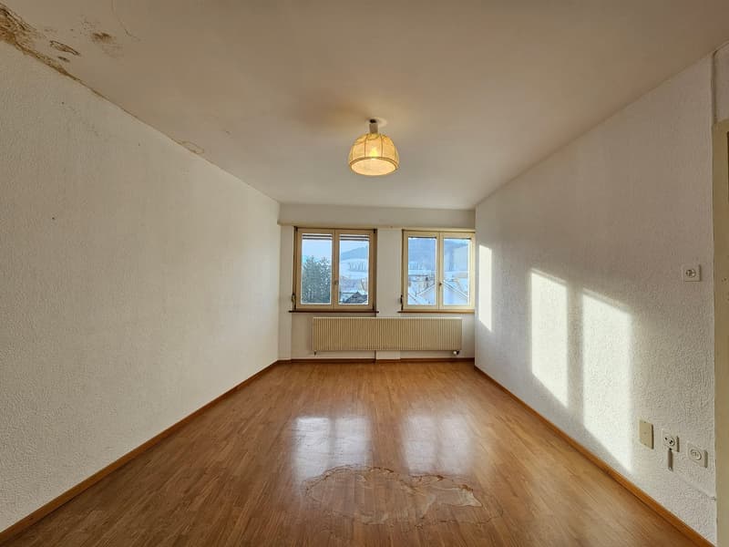 Empty room with wooden floor, two windows, white walls, central lamp, white outlets.