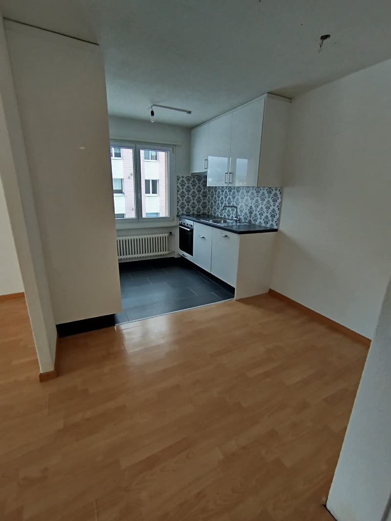 Small, empty kitchen with wooden floors, white kitchen units, dark countertops, white radiator, and window