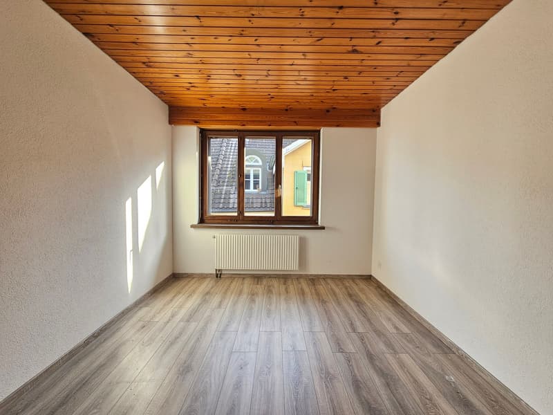 An empty room with wooden floors, white walls, a wooden ceiling, a window, and a white radiator. The window has a view of a house with a yellow facade and green shutters. The room has a sense of minimalism.