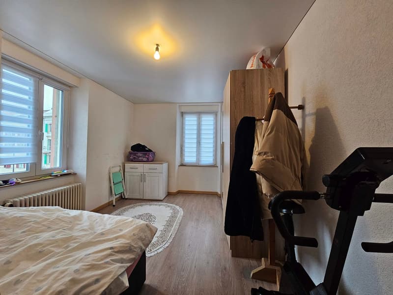 Bedroom with bed, white wardrobe, small wooden cabinet, two windows, beige carpet, light, coat hanging on a rack, and a piece of exercise equipment.