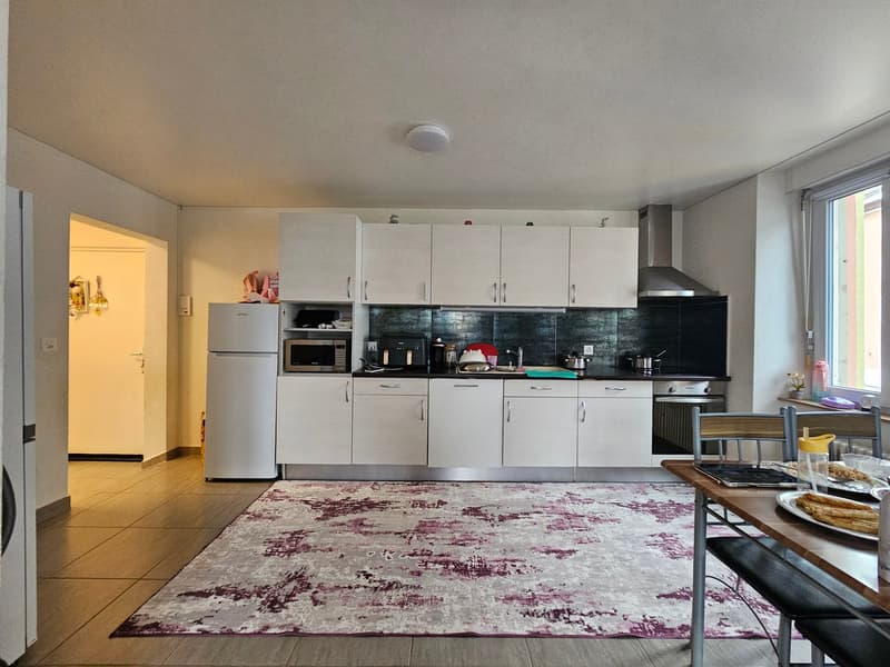 White kitchen with tile floor, microwave, refrigerator, countertop, sink, stove, cabinets, rug, and dining area with a table and chairs.