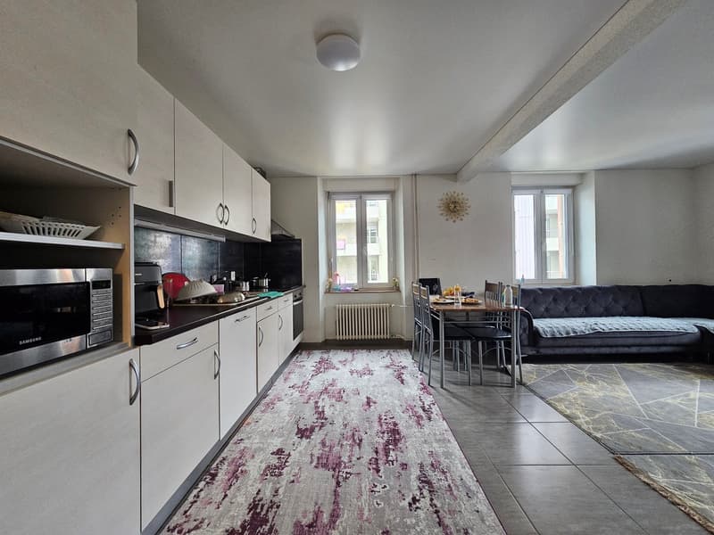 modern kitchen with white cabinets, gray tiles, and white countertops, equipped with a microwave, refrigerator, oven, and a dining area with a table and chairs, a cozy living room with a couch, wooden dining table, and white radiator, and two large window