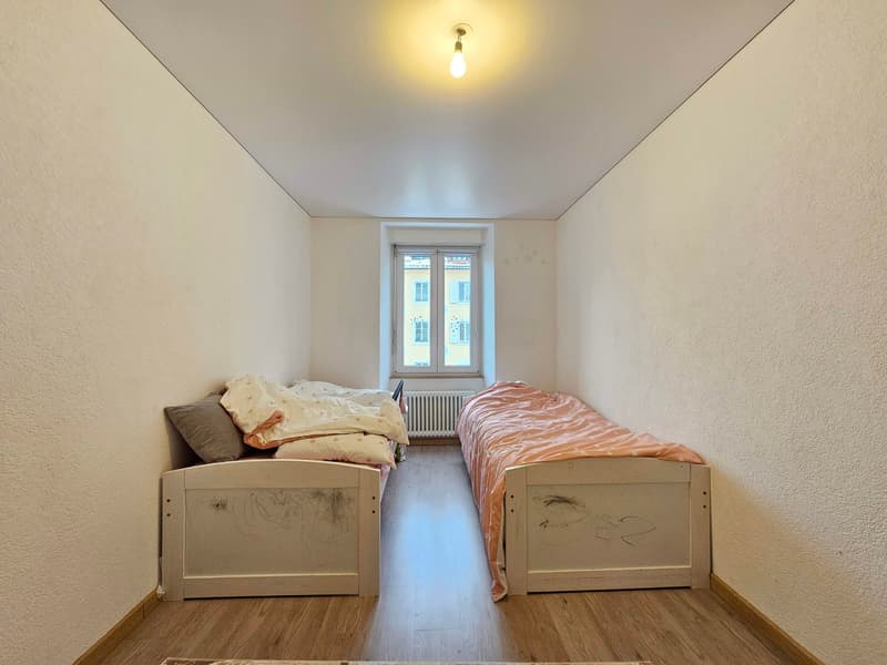 Small room with two beds, white walls, wooden floor, light fixture, window, radiator