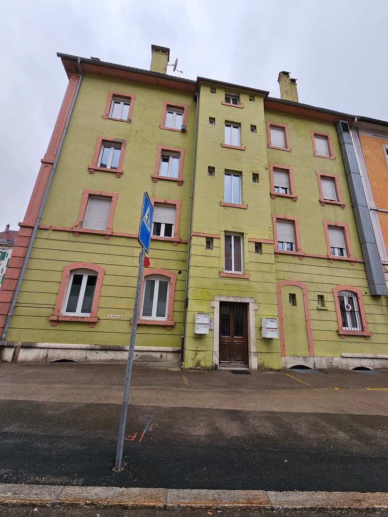 An old green apartment building with several windows, a small balcony on the second floor, and a door leading to the street.