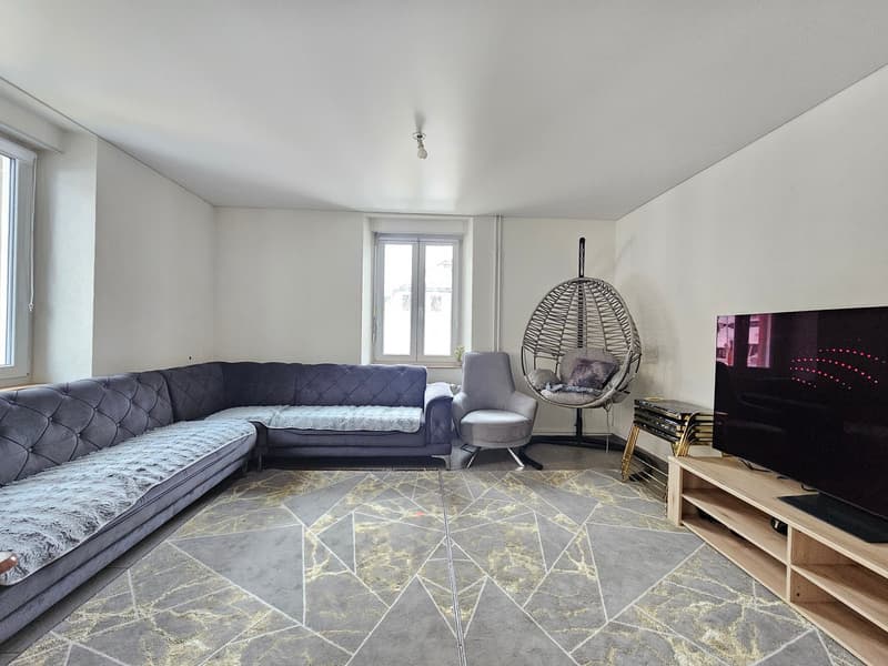 Large living room with grey velvet sofa, light walls, modern design floor lamp, hanging chair, TV, and white tile floor.