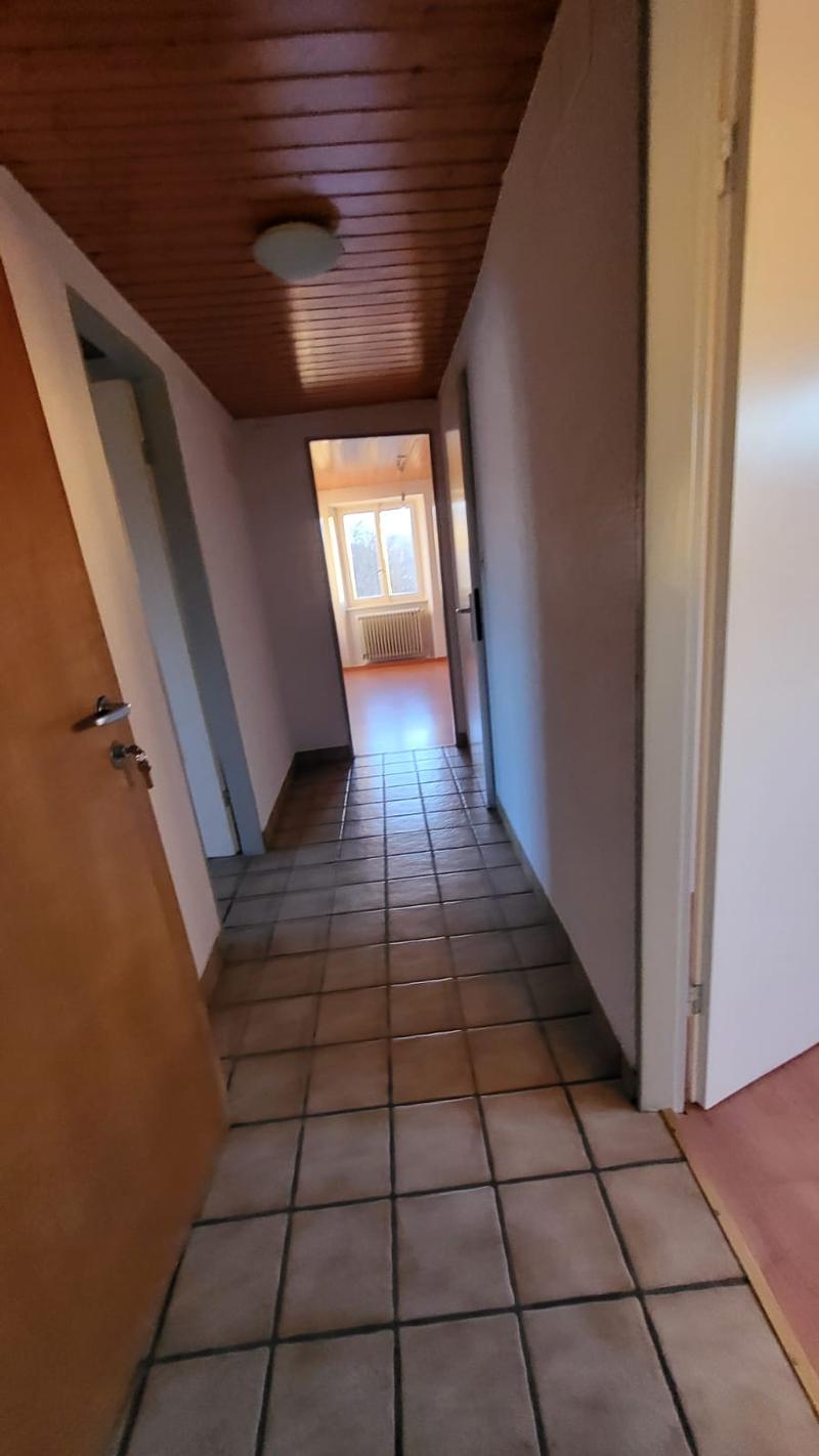 Tiled floor, ceiling with ceiling lights, walls are painted white, radiator on one wall, doorway in the middle, door on the left side.