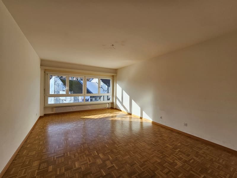 Empty room with white walls, wooden parquet floor, large windows with view of cityscape.