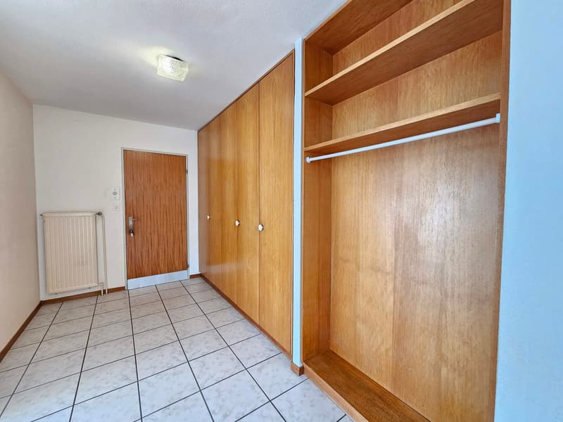 Empty hallway with white tiles, large wooden wardrobes, radiator, wooden door, open wardrobe, shelf, metal rod.