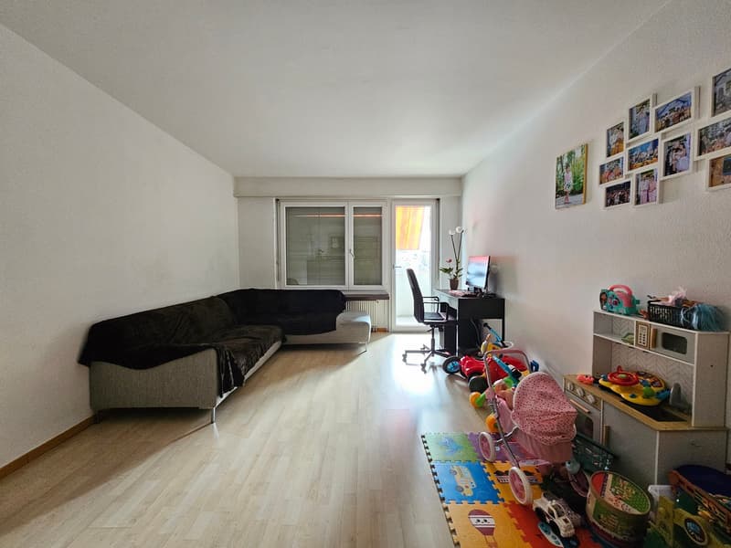 ample living room, L-shaped sofa, desk with computer and chair, glass door leading to a balcony, wall decoration with photos, floor covered with children's toys