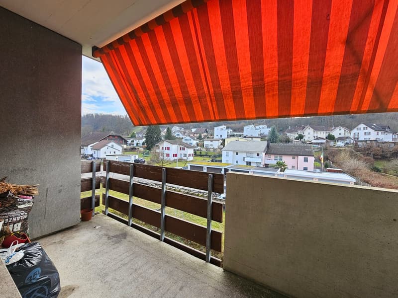 A balcony with a wooden railing and a red awning overlooking a village.