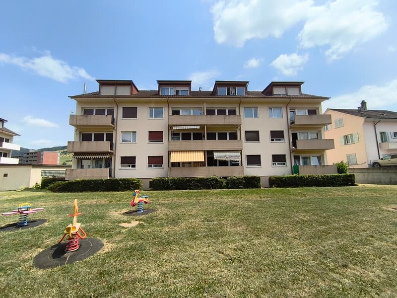 A multi-story apartment building with balconies, surrounded by a well-maintained lawn and play equipment for children.