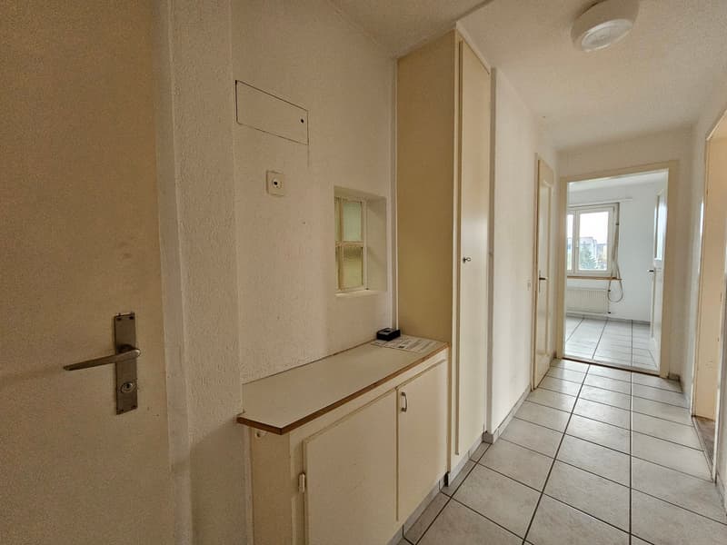 Empty hallway with tiled floor, white walls, a small counter with cabinets, a power outlet, and a small window.