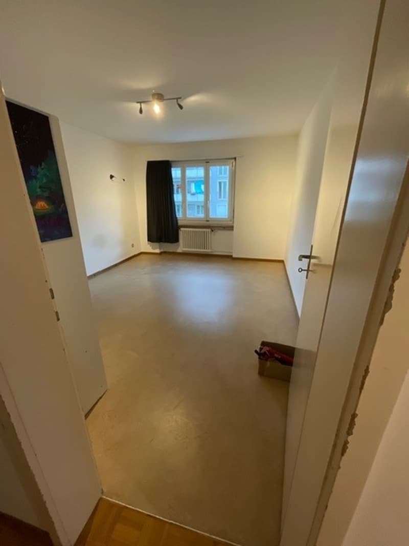 Empty room with white walls, wooden floor, window with black curtains, radiator, ceiling lights, and a small box on the floor.